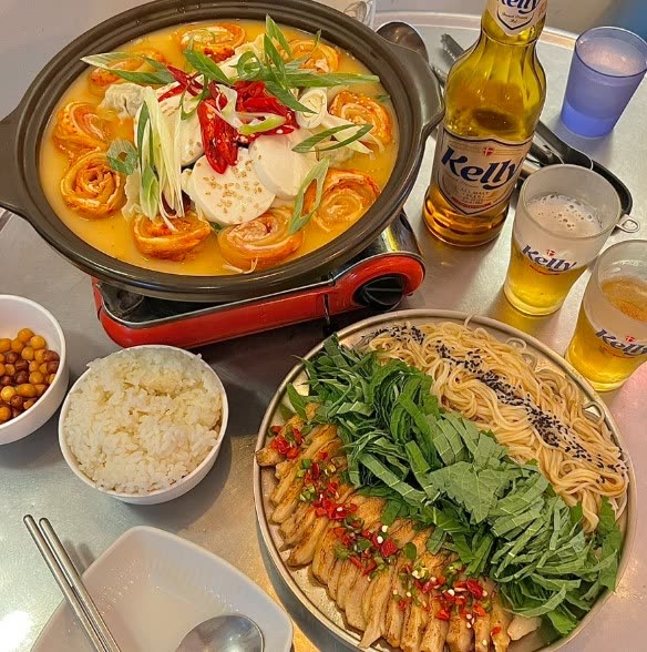 두부와 새우가 든 전골 냄비가 가스버너 위에서 끓고, 수육과 소면·깻잎을 담은 접시, 공기밥, 맥주 두 잔이 한 상에 차려져 있다