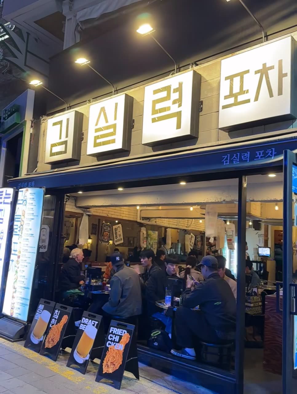 밤의 김실력포차 외관 — 네 개의 조명 간판에 '김 실 력 포 차'가 한 글자씩 켜져 있고, 통유리 안쪽으로 손님들이 앉아 있는 이태원 술집